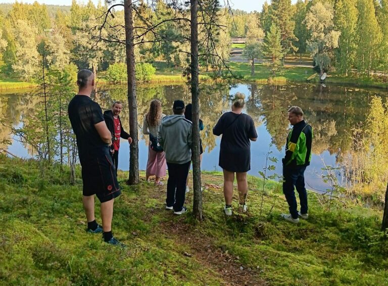 Seitsemän ihmistä järven rannalla, sammaleinen maa. Kasvot eivät näy.